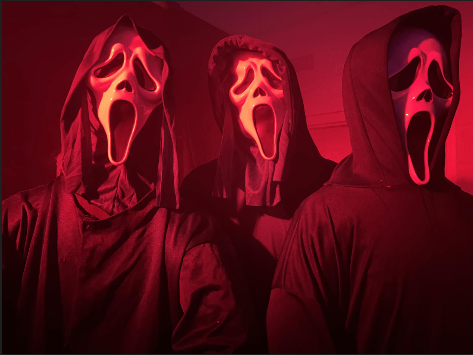 Three people wearing black hooded cloaks and ghost face masks stand in a dimly lit room illuminated by red light, evoking a mysterious and eerie atmosphere